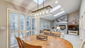 A dining area with a wooden table and chairs, a hanging light fixture, and a view of the outside through large windows.