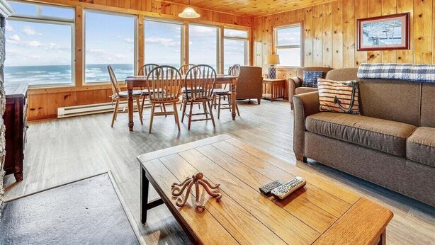 A wooden cabin interior with a dining area, sofa, and large windows overlooking the ocean.