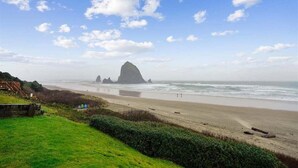 A beach with a large rock formation in the distance, a grassy area in the foreground, and a clear sky.