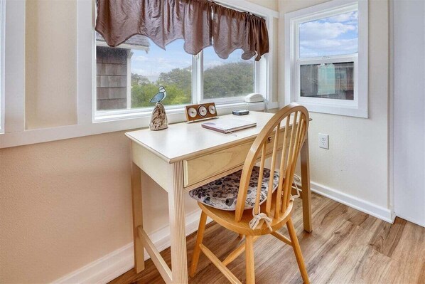 Une pièce comprenant un bureau en bois, une chaise, une fenêtre avec des rideaux, une petite table ornée d’objets décoratifs et une vue sur la verdure à l’extérieur.