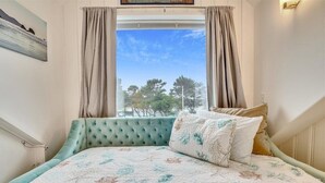 A bedroom with a bed, curtains, a window with a view of trees, and a painting on the wall. - Sandy Owl - Close Beach Access! (Cannon Beach)