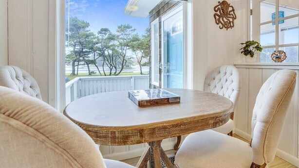 A dining area with a round wooden table, white upholstered chairs, and a view of trees through a glass door. - Sandy Owl - Close Beach Access! (Cannon Beach)