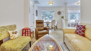 A living room with a brown leather armchair, a yellow sofa, and a glass coffee table. There is a red stool and a white chair in the background. - Sandy Owl - Close Beach Access! (Cannon Beach)