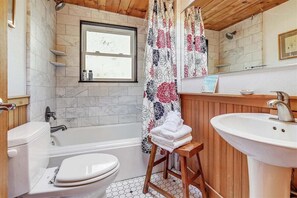 A bathroom with a white bathtub, a white toilet, a wooden stool with towels, a white sink, and a window with a view. - Salal Bungalow - Close to Beach & Downtown! (Cannon Beach)