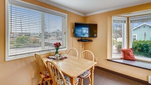 A dining area with a table and chairs, a television mounted on the wall, and large windows with blinds. - Sandcastle Inn #601 - Downtown Apartment! (Cannon Beach)