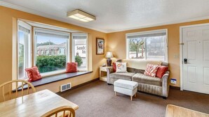 A cozy living room with a sofa, ottoman, and a dining table. Large windows offer a view of the outside. - Sandcastle Inn #601 - Downtown Apartment! (Cannon Beach)