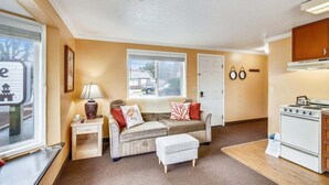 A compact living space with a sofa, ottoman, a small table with a lamp, a window with blinds, a door, and a kitchen area with a stove. - Sandcastle Inn #601 - Downtown Apartment! (Cannon Beach)