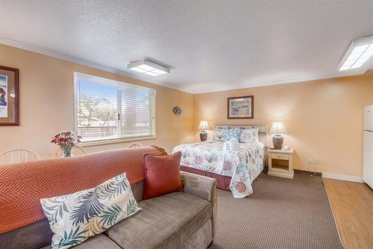 A bedroom with a bed, a sofa, a nightstand, a lamp, and a window with blinds.