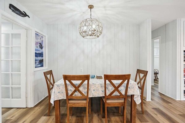 A dining area with a round table, four wooden chairs, a chandelier, and a white wall with a framed picture.