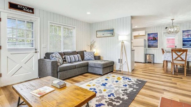 Ein Wohnzimmer mit einer grauen Eckcouch, einem Holztisch und einem gemusterten Teppich. Es gibt einen Essbereich mit Tisch und Stühlen, und im Hintergrund eine Küche.