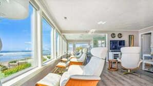 A modern room with a long bench seating area, large windows with a view of the ocean, and a television on the wall. - Schilling Beach House (Arch Cape)