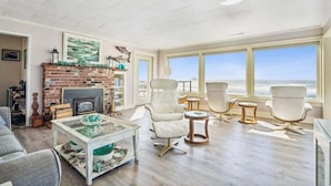 A spacious living room with a fireplace, white leather chairs, a glass coffee table, and large windows overlooking the ocean. - Schilling Beach House (Arch Cape)