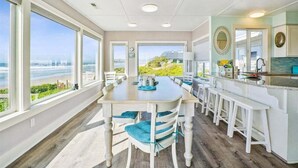 A dining area with a view of the ocean and hills, featuring a large table and white bar stools. - Schilling Beach House (Arch Cape)