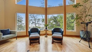 A spacious room with large windows, two leather armchairs, a small table with a plant, and a tripod with a camera. - Santo Cristo - Ocean View! (Arch Cape)