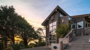 A modern wooden house with a staircase leading to the entrance, surrounded by trees and a gravel driveway. - Santo Cristo - Ocean View! (Arch Cape)