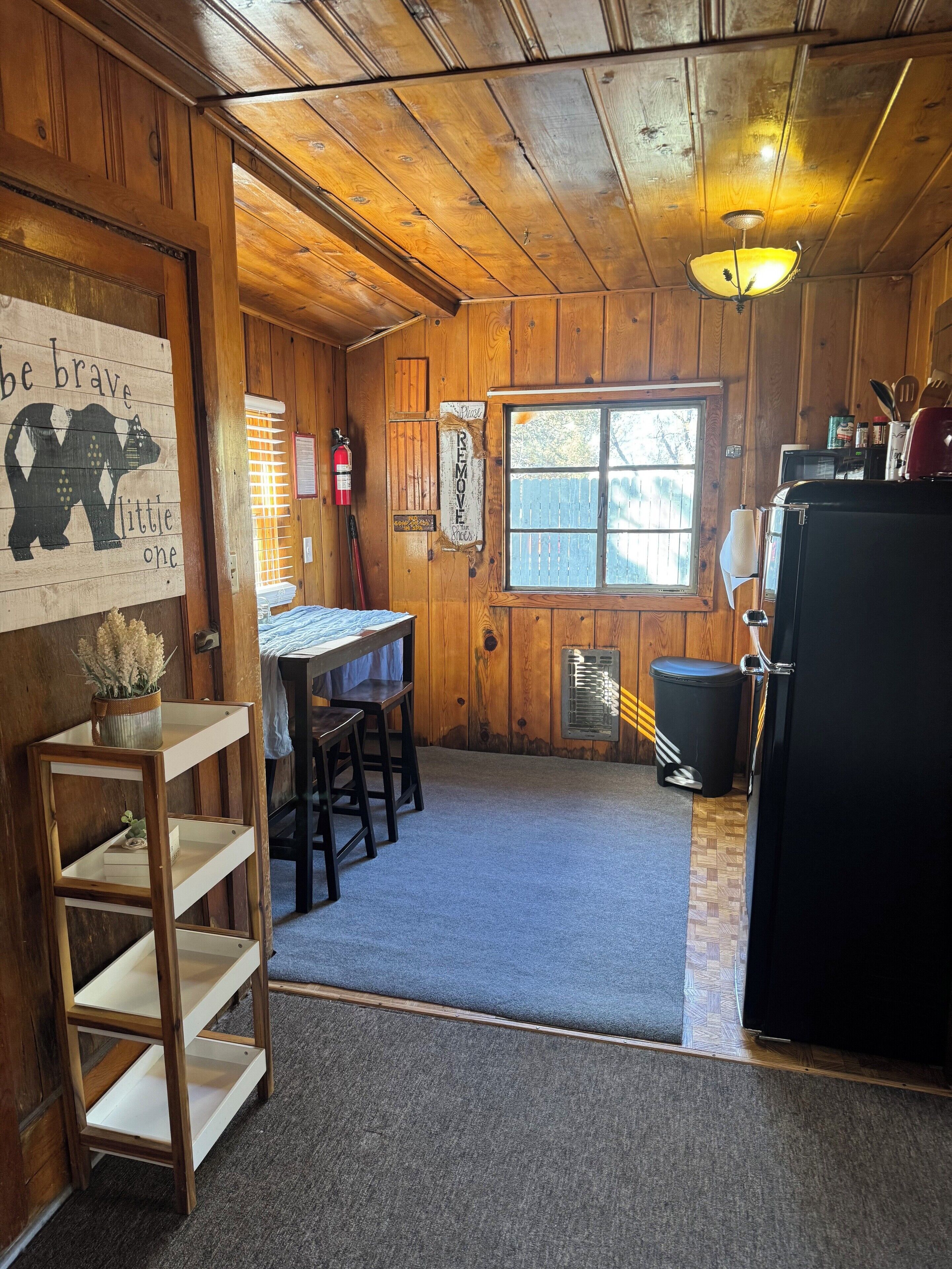 A rustic cabin interior with wooden walls, a kitchen area with a refrigerator, a sink, and a window.