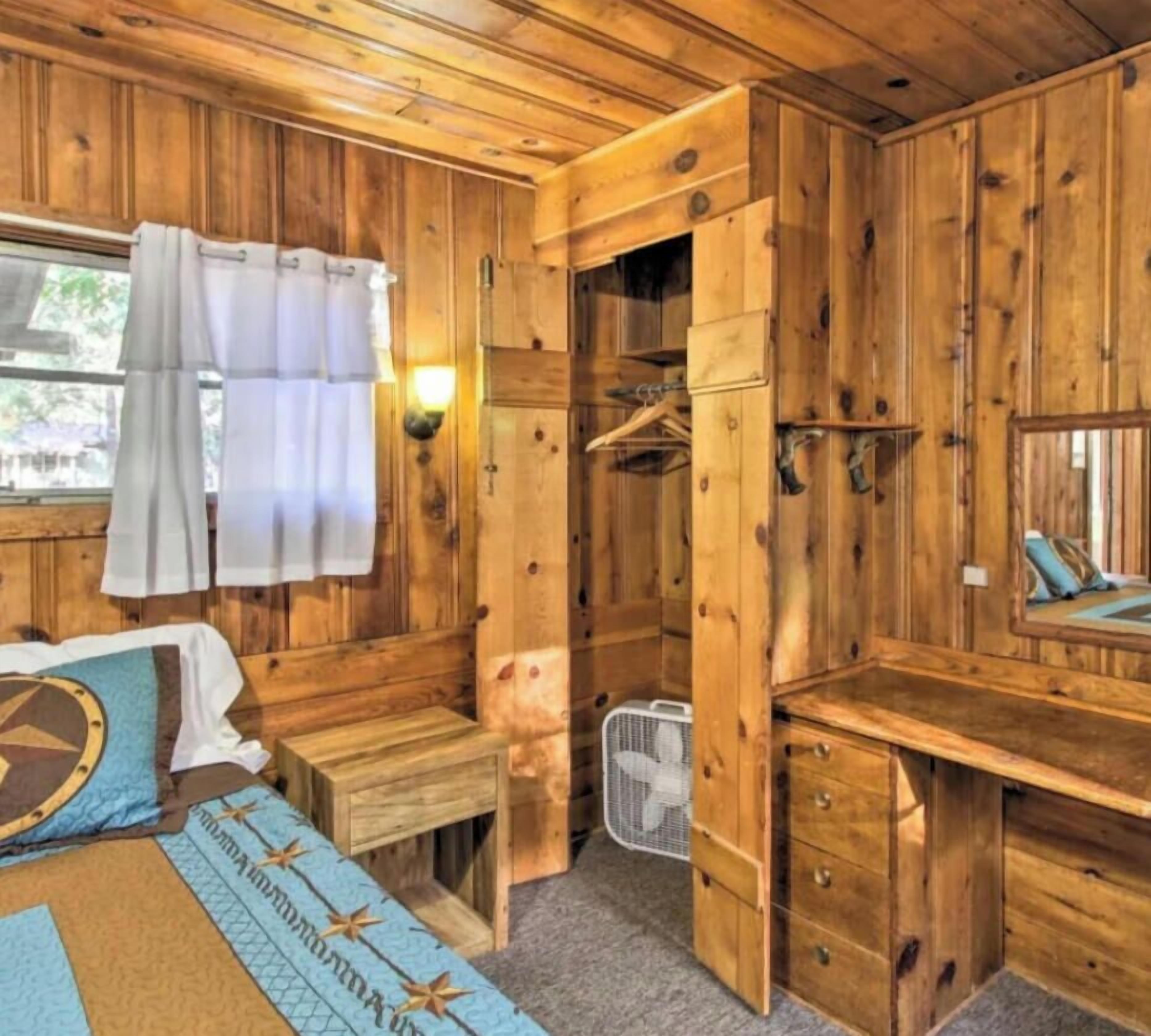 A wooden room with a bed, a desk, a hanging coat rack, and a window with curtains.