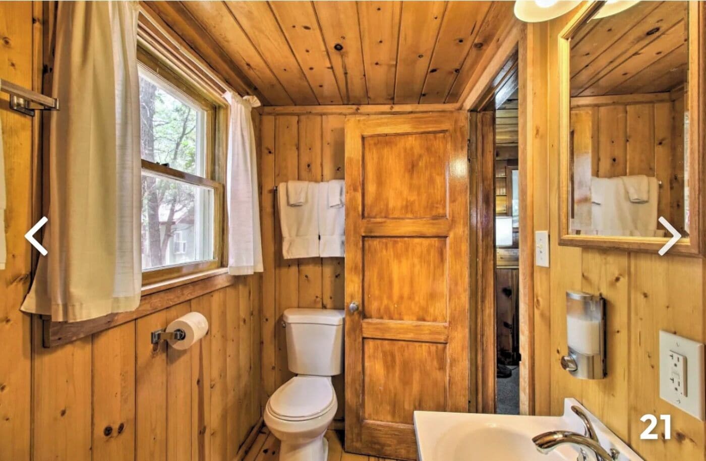 A wooden bathroom with a toilet, sink, and window with curtains.
