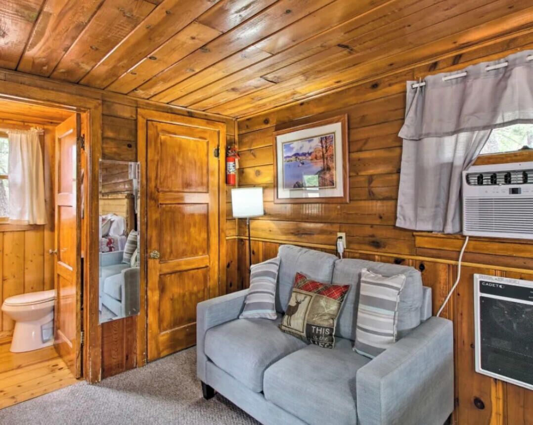 Ein Blockhaus-Interieur mit Holzbalkendecke, einer Couch mit Kissen, einem gerahmten Bild an der Wand und einem Fenster mit Vorhängen.
