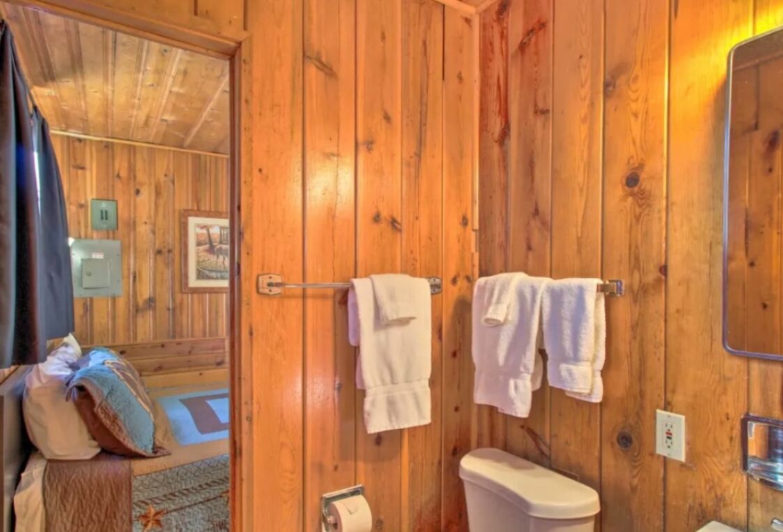A wooden-paneled bathroom with a toilet, two towels hanging on a rack, and a visible electrical outlet.