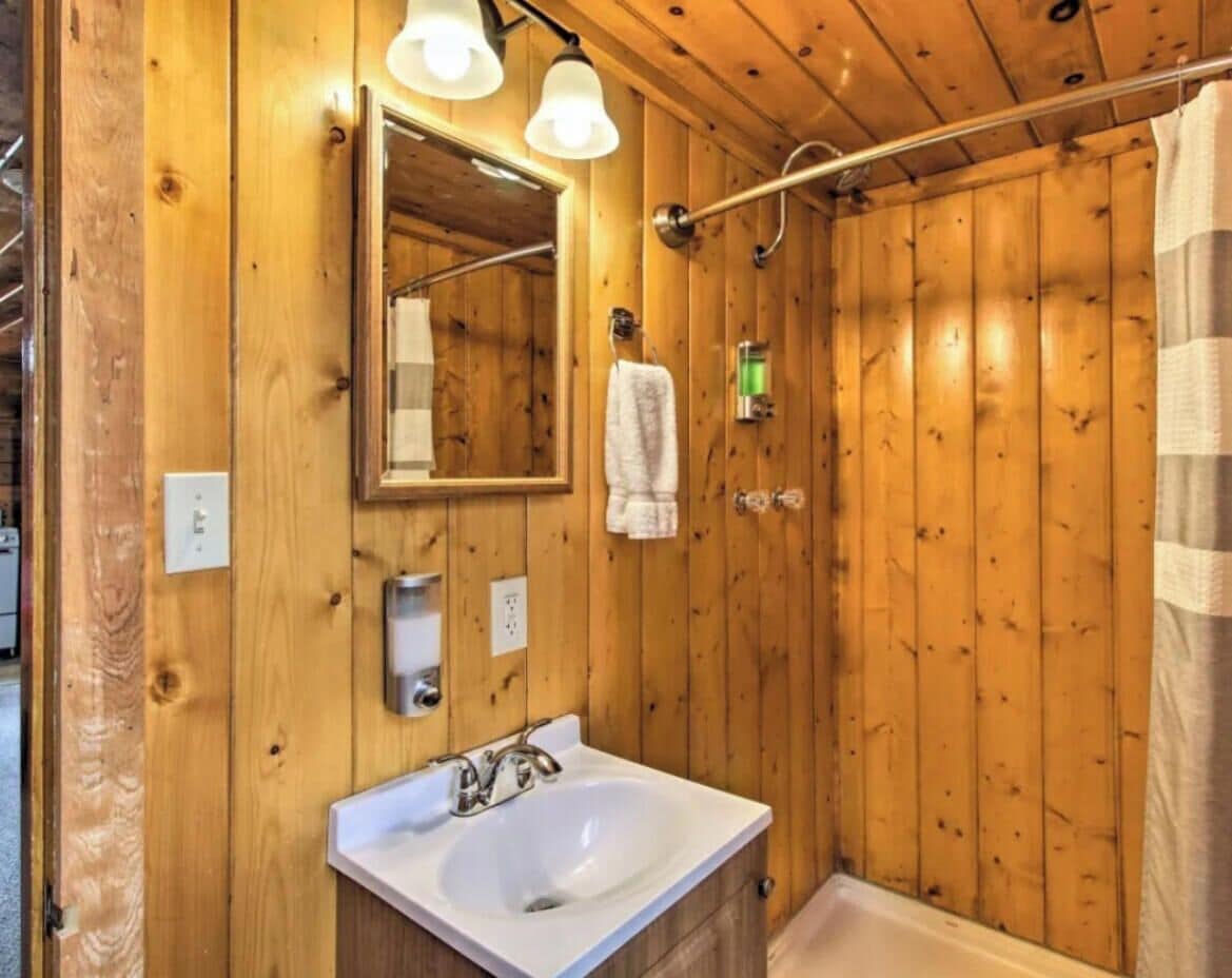 Une salle de bain aux murs lambrissés, équipée d’un lavabo, d’un miroir, d’un porte-serviettes et d’un rideau de douche.