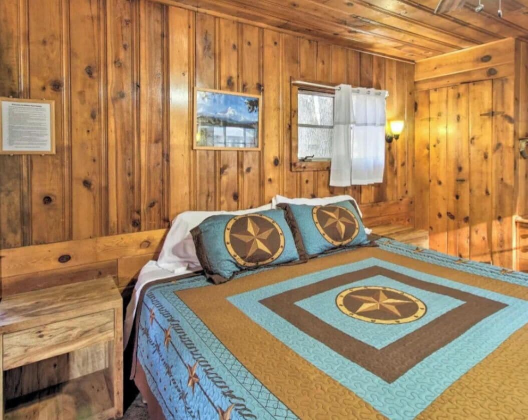 A wooden-paneled room with a bed, a nightstand, a window with curtains, and a wall-mounted light.