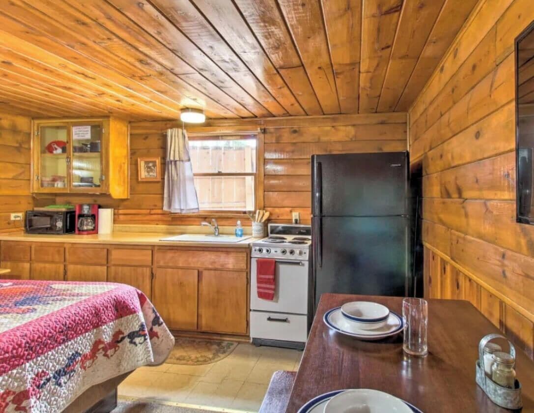 A wooden cabin interior with a kitchen area, a dining table, and a bed.