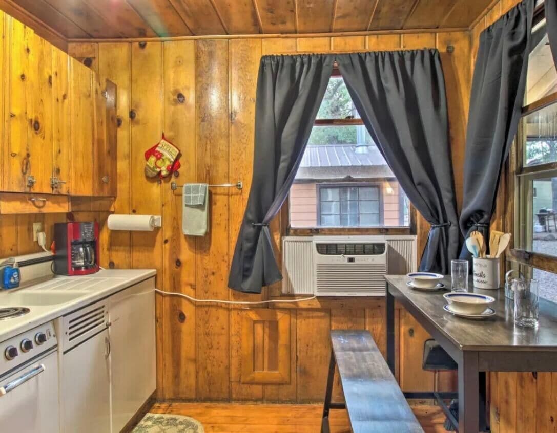 A compact kitchen with wooden walls, a window with curtains, a small table, and a bench.