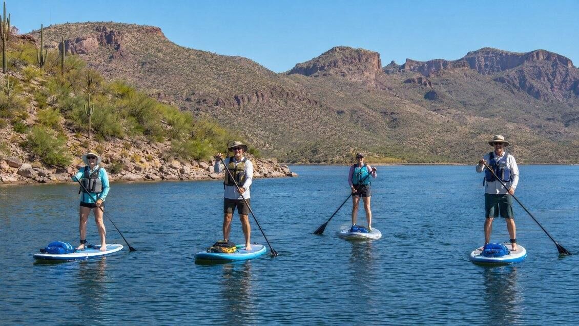 Four individuals stand-up paddleboarding on a calm body of water with a mountainous landscape in the background.