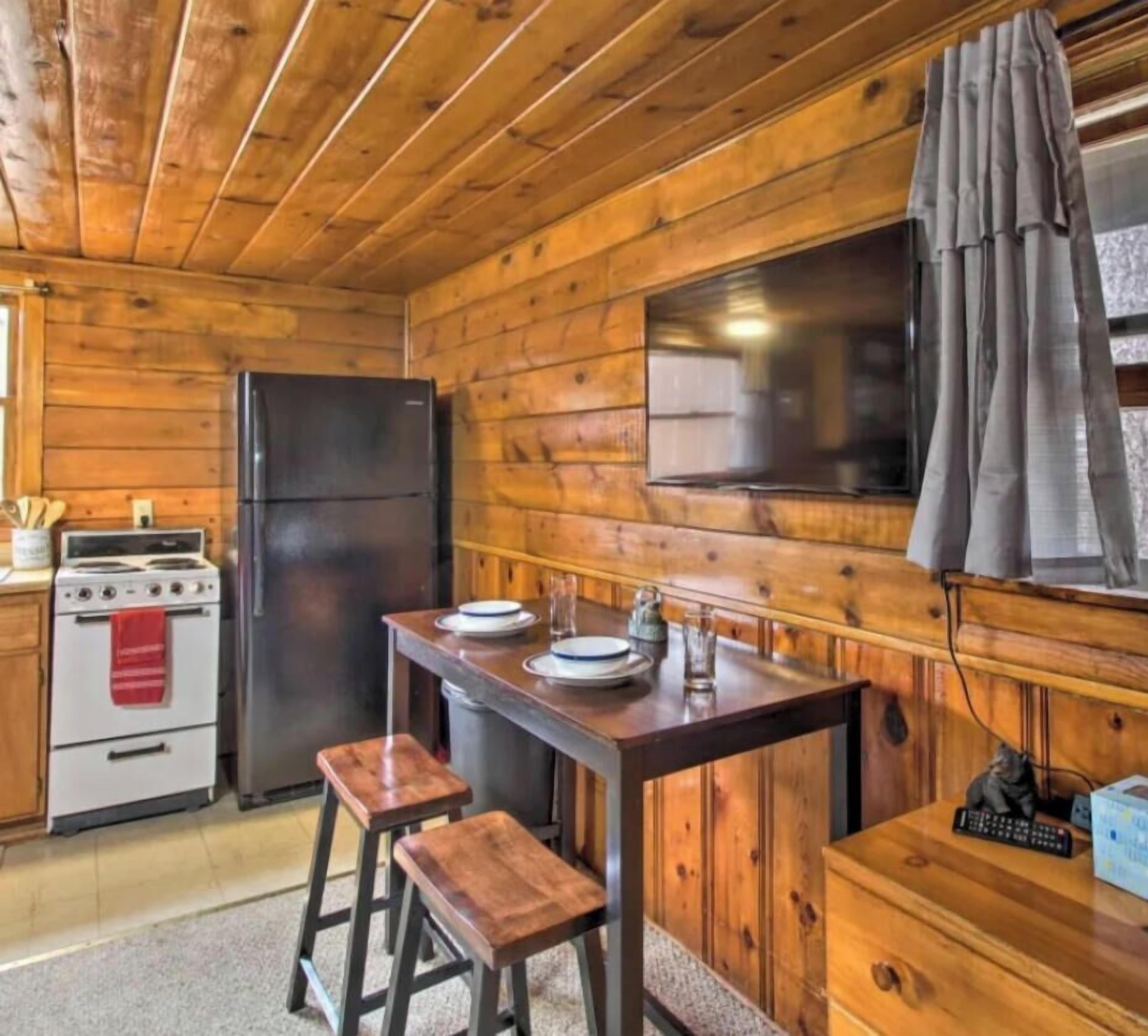Interior de una cabaña de madera con una cocina que incluye estufa, refrigerador y una mesa de madera con taburetes.