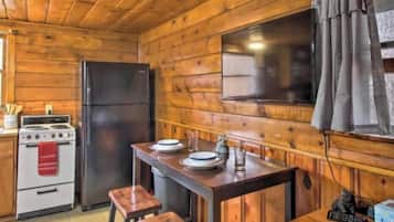 Interior de una cabaña de madera con una cocina que incluye estufa, refrigerador y una mesa de madera con taburetes.