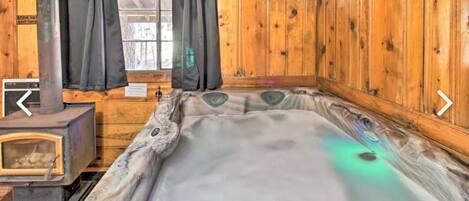 A wooden cabin interior with a hot tub, a wood-burning stove, and a window with curtains.