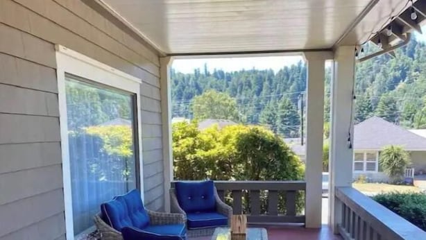 Een overdekt terras met rieten meubels en een glazen tafel, met uitzicht op een bebost gebied.