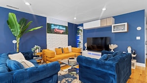 A modern living room with a blue sofa set, a yellow sofa, and a coffee table. There is a TV mounted on the wall, a large plant, and a neon sign that reads "Sweet Home Chicago". - Luxe Duplex - Private Gym -Huge Backyard -Arcade- King Beds - Garage-EV Charger (Chicago)