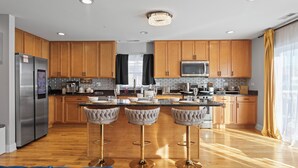 A modern kitchen with wooden cabinets, stainless steel appliances, and a central island with bar stools. - Luxe Duplex - Private Gym -Huge Backyard -Arcade- King Beds - Garage-EV Charger (Chicago)