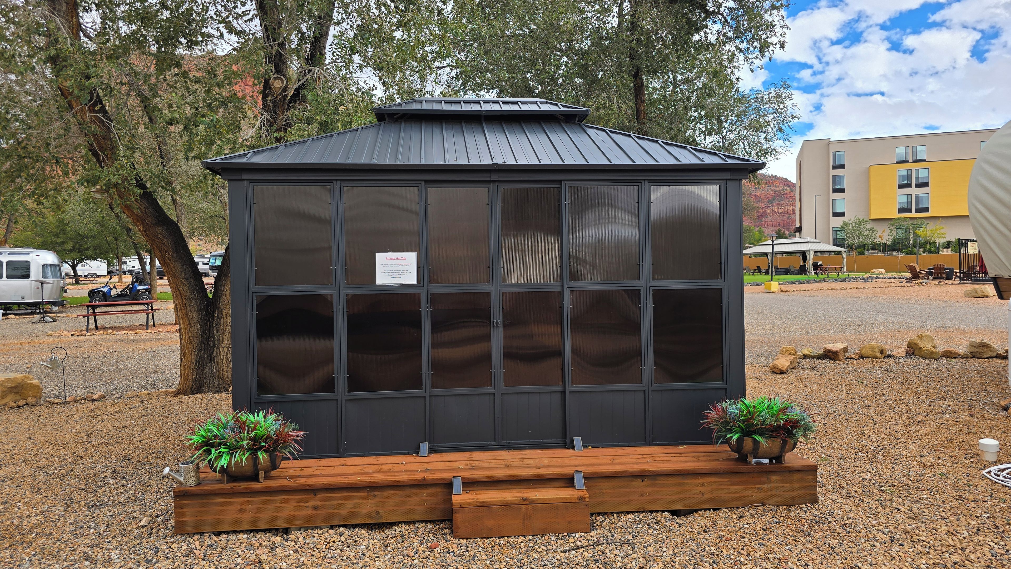 A small, dark-colored, glass-enclosed structure with a metal roof, surrounded by gravel, with two potted plants on wooden stands.