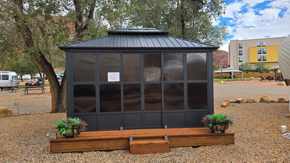 A small, dark-colored, glass-enclosed structure with a metal roof, surrounded by gravel, with two potted plants on wooden stands.