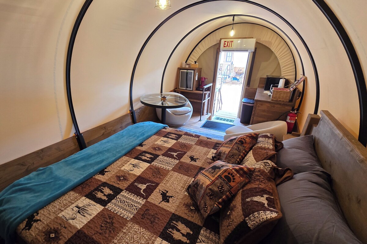 A cozy room with a bed, a small table, and a view of the outside through an arched window.