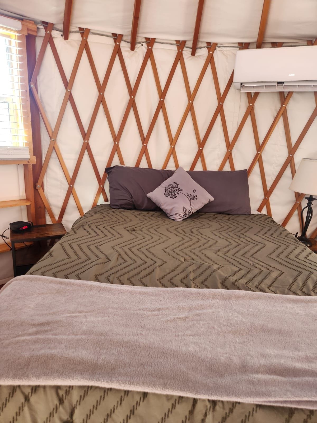 Una cama con colcha estampada, almohadas y manta en una habitación tipo tienda con pared de celosía de madera y un equipo de aire acondicionado.