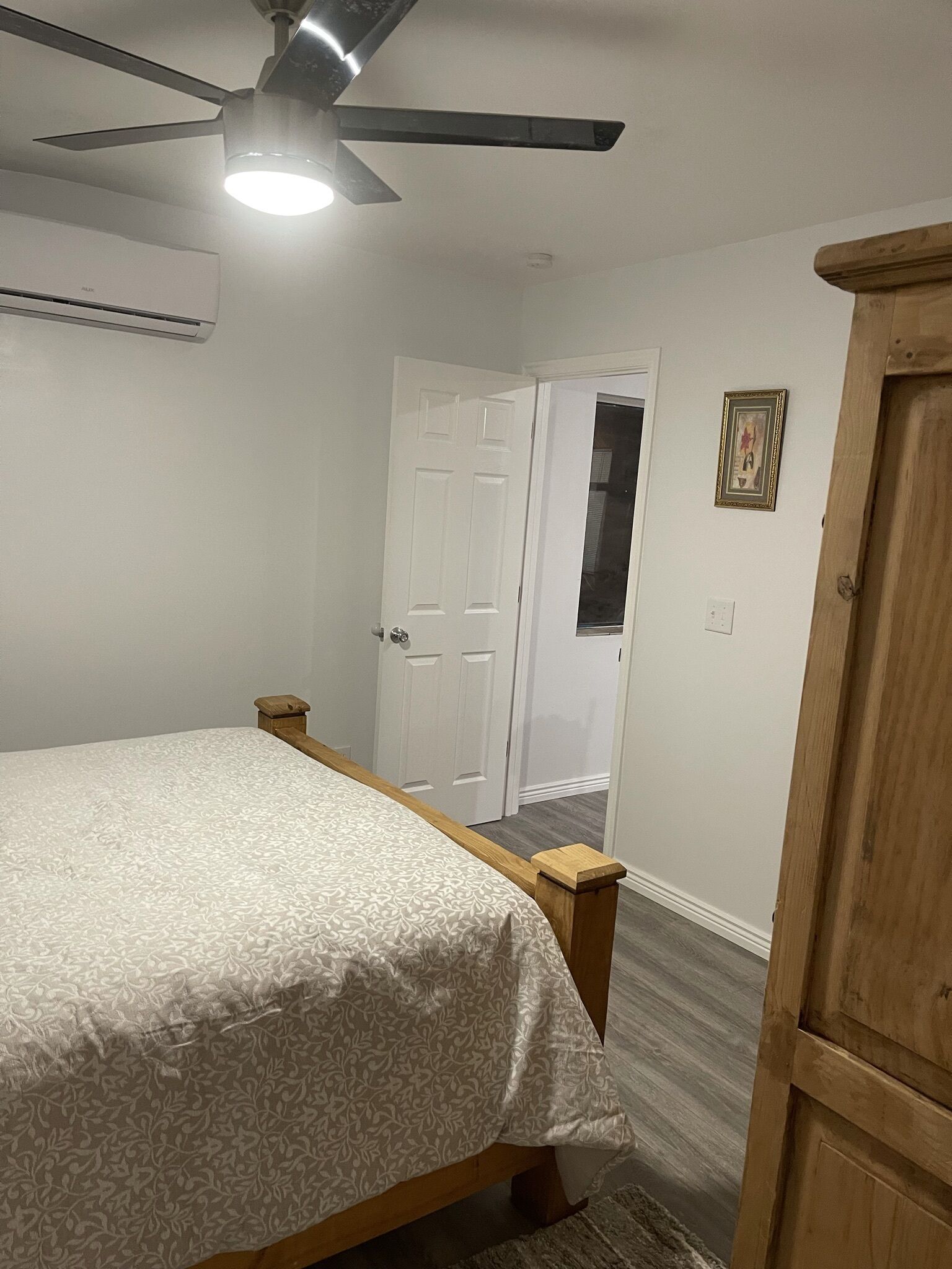 Un dormitorio con cama, ventilador de techo, equipo de aire acondicionado, armario de madera y una puerta que da a otra habitación.