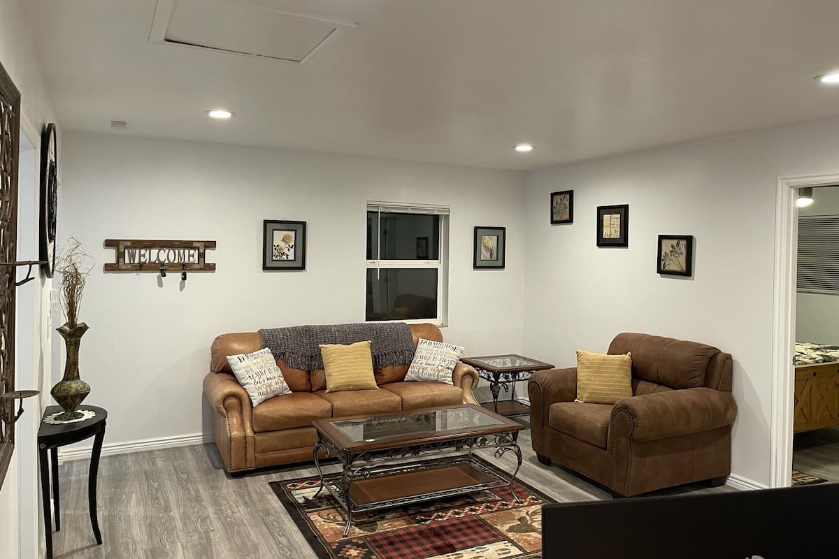 A living room with a sofa, armchair, coffee table, and decorative pillows. There is a window, wall art, and a welcome sign.