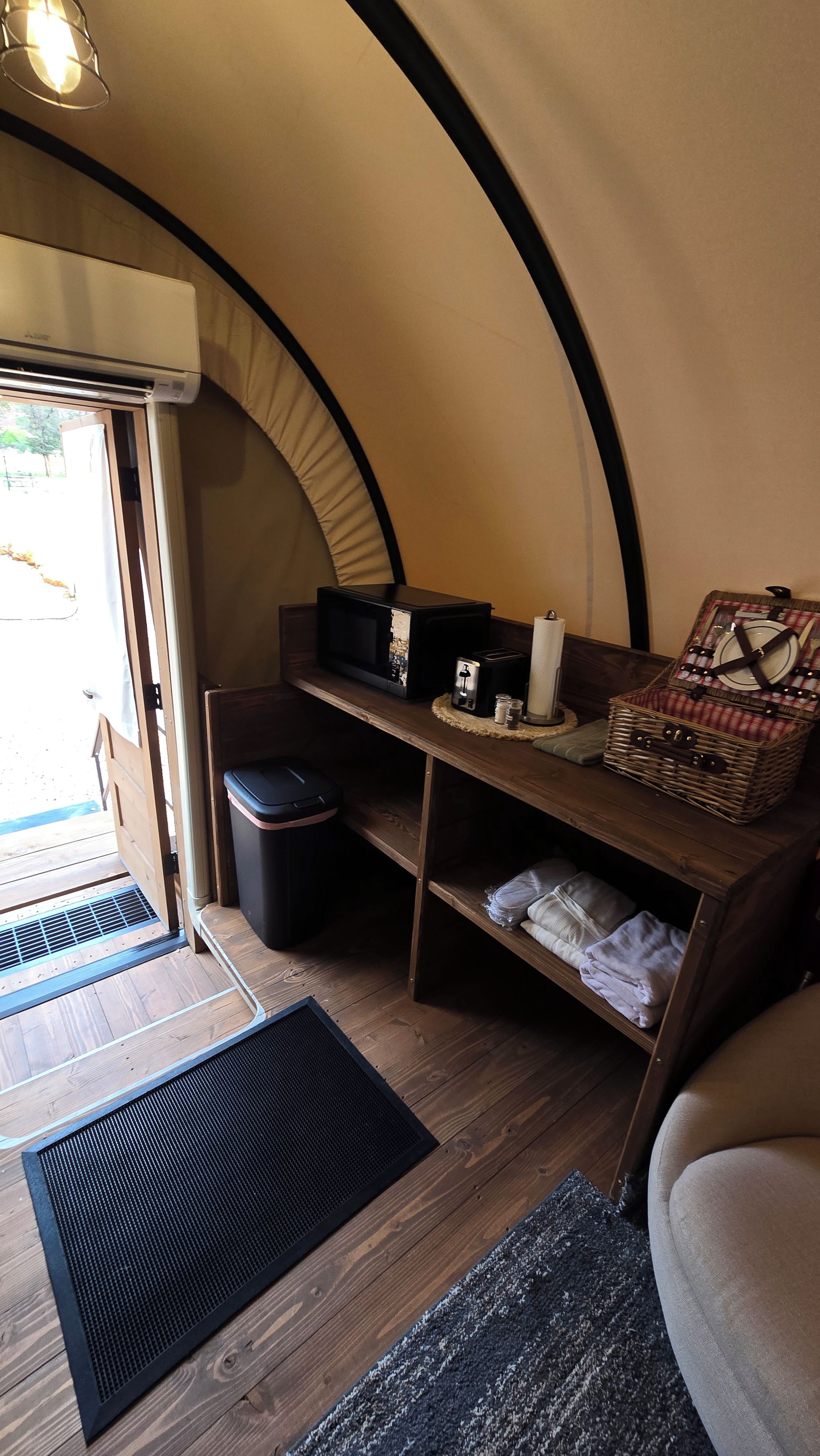 A compact living space with a kitchenette, a small trash bin, a bench, and a storage chest.