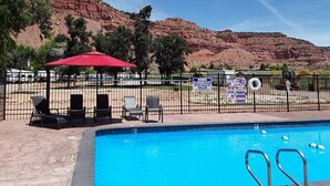 A swimming pool with lounge chairs and a red umbrella, surrounded by a fenced area with desert hills in the background. - Crazy Horse Rv Resort (Kanab)