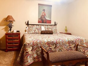 A bedroom with a bed, a nightstand, a lamp, a chair, and a wall-mounted artwork. - MJ’S BUNKHOUSE #4 (Pawhuska)