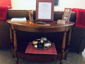 Una mesita de madera con un letrero de bienvenida en marco, un pequeño florero y un cuenco decorativo con adornos.