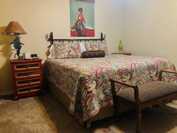 Un dormitorio con cama, mesita de noche, lámpara, silla y un cuadro en la pared.