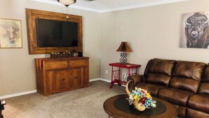 A living room with a brown leather sofa, a wooden cabinet, a round coffee table with a floral arrangement, a television on a wooden stand, and a red lamp.