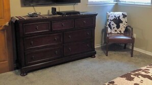 A bedroom with a dark wooden dresser, a flat-screen TV mounted above it, a cow-print chair, and a bed with a cow-print bedspread.