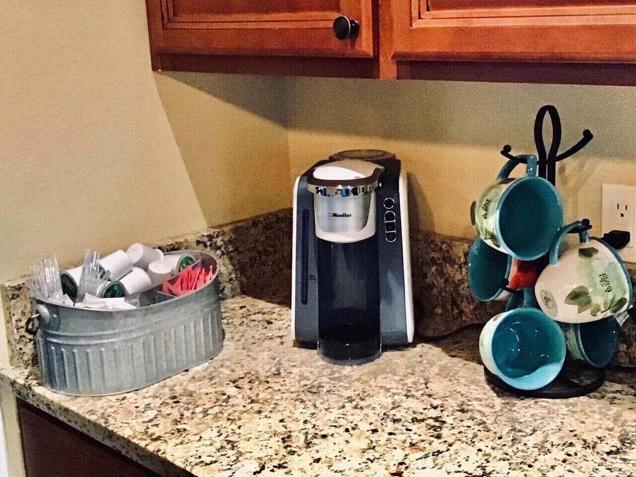 A kitchen counter with a coffee maker, a metal container with utensils, and a set of blue cups.