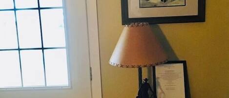 A room with a door, a window, a framed picture on the wall, a lamp, and a small table with a book and a certificate.